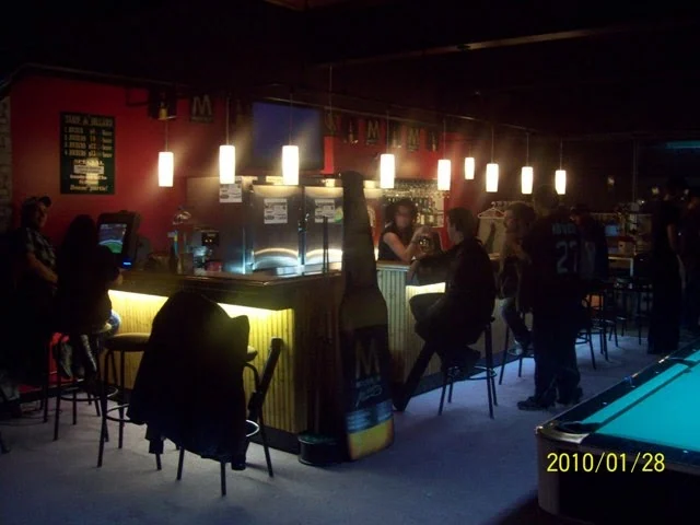 Le bar au fond de la salle, éclairé par une rangée de suspensions, avec des clients assis sur des tabourets.