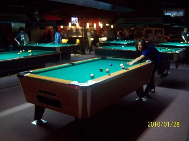 Plusieurs tables de billard éclairées dans la salle ; une joueuse s’apprête à frapper la bille.